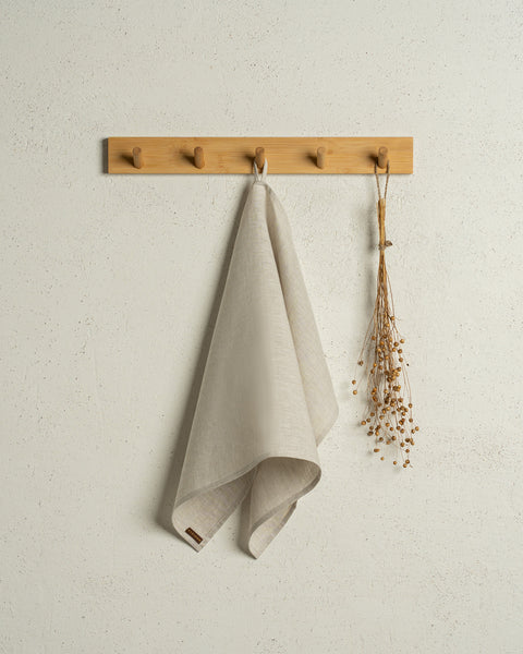 The menique linen kitchen dish towel in the color of natural hanging on a wooden rack on a wall.
