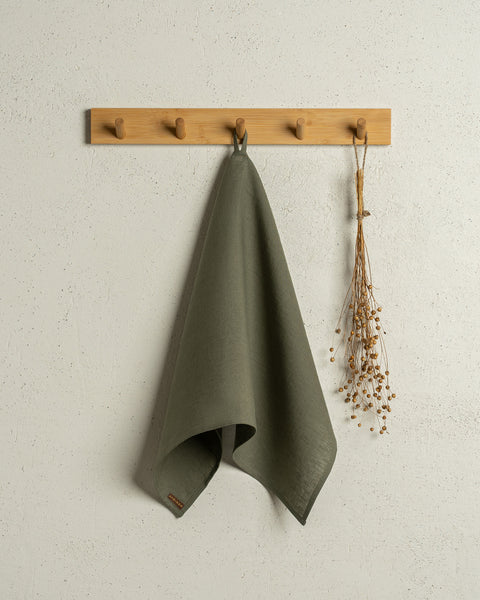 The menique linen kitchen dish towel in the color of stone green hanging on a wooden rack on a wall.