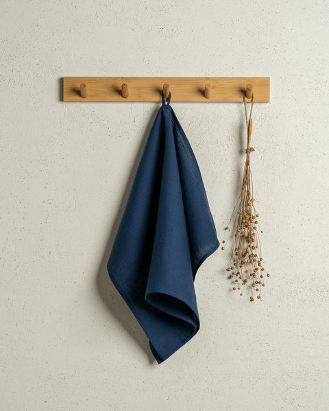 The menique linen kitchen dish towel in the color of storm blue hanging on a wooden rack on a wall.
