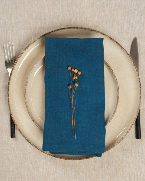 The menique linen napkin in cobalt blue color, put on top of two stacked plates . There are cutlery items by the sides of the plates. The background is a natural color linen tablecloth.