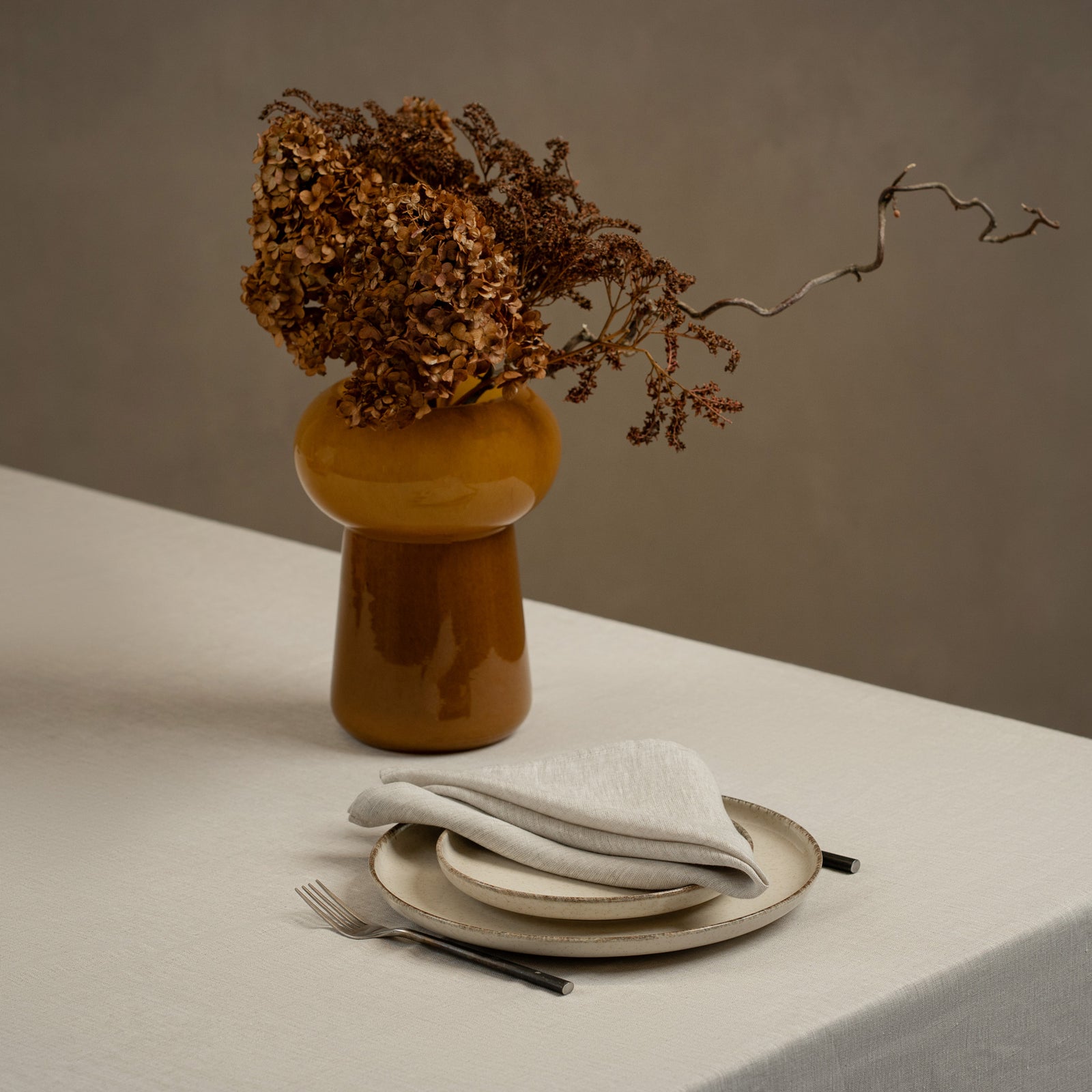 A tabletop covered with a linen tablecloth, with a vase and a single plate set on top of it. By the sides of the plates there are cutlery items, and there is a linen napkin in cobalt blue color on top.
