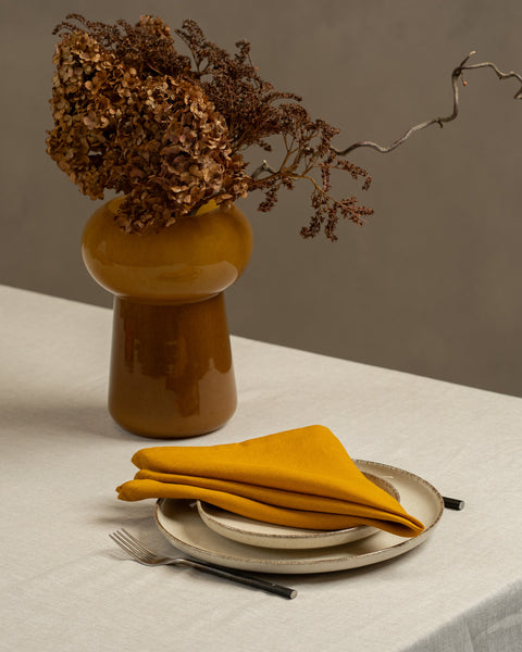 A table covered with a linen tablecloth, with a vase and plates on top of it. On top of the stacked plates is a Linen Napkin in Spicy Yellow color.
