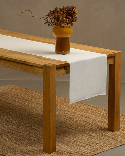 The pure white linen table runner on top of a wooden table, captured in a close up picture from the corner of the table. 