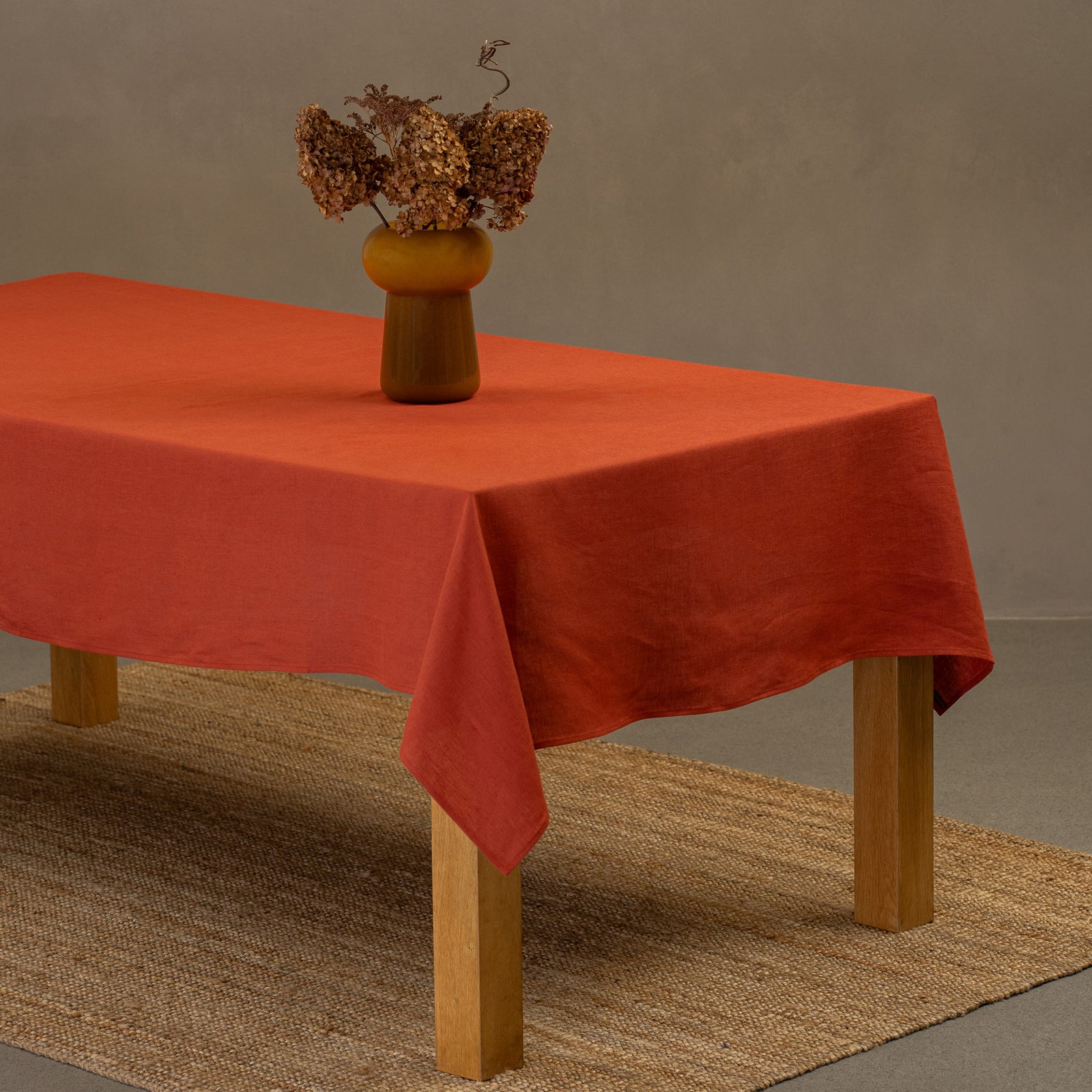 Het menique linnen tafelkleed in steengroene kleur op een houten tafel. Op het tafelkleed staat een vaas gemaakt van natuurlijke stroken en een kleien beker. Onder de tafel ligt een kleed.