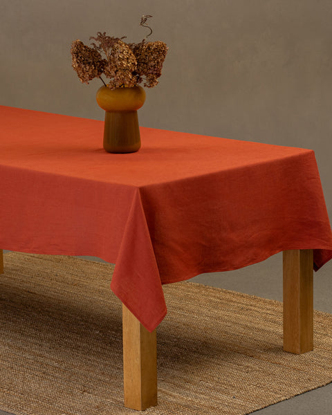 Het menique tafelkleed van linnen in kaneelrood op een houten tafel. Op het tafelkleed staat een vaas gemaakt van natuurlijke stroken en een kleikopje. Onder de tafel ligt een kleed.