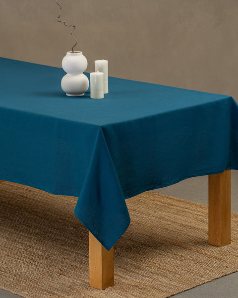 Het menique cobaltblauwe linnen tafelkleed op een houten tafel. Op het tafelkleed staat een vaas en kaarsen. Onder de tafel ligt een kleed.