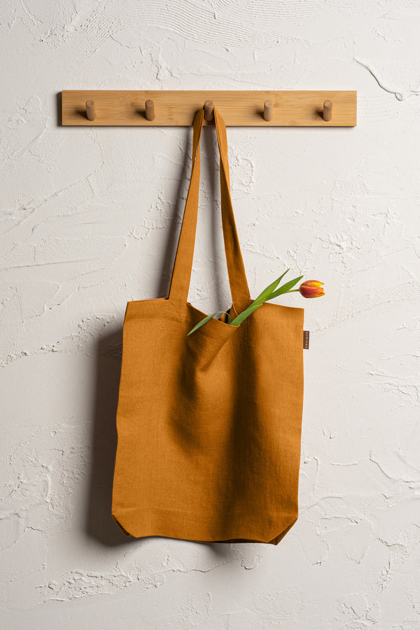 Menique spicy yellow durable linen tote bag hangs from a wooden rack with four pegs mounted on a textured off-white wall. 