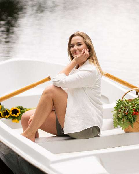 A woman relaxes inside a small white rowboat on calm water, gently resting her chin on her hand. She wears a white shirt and olive green shorts, sitting near a basket of fresh greens and bright sunflowers.