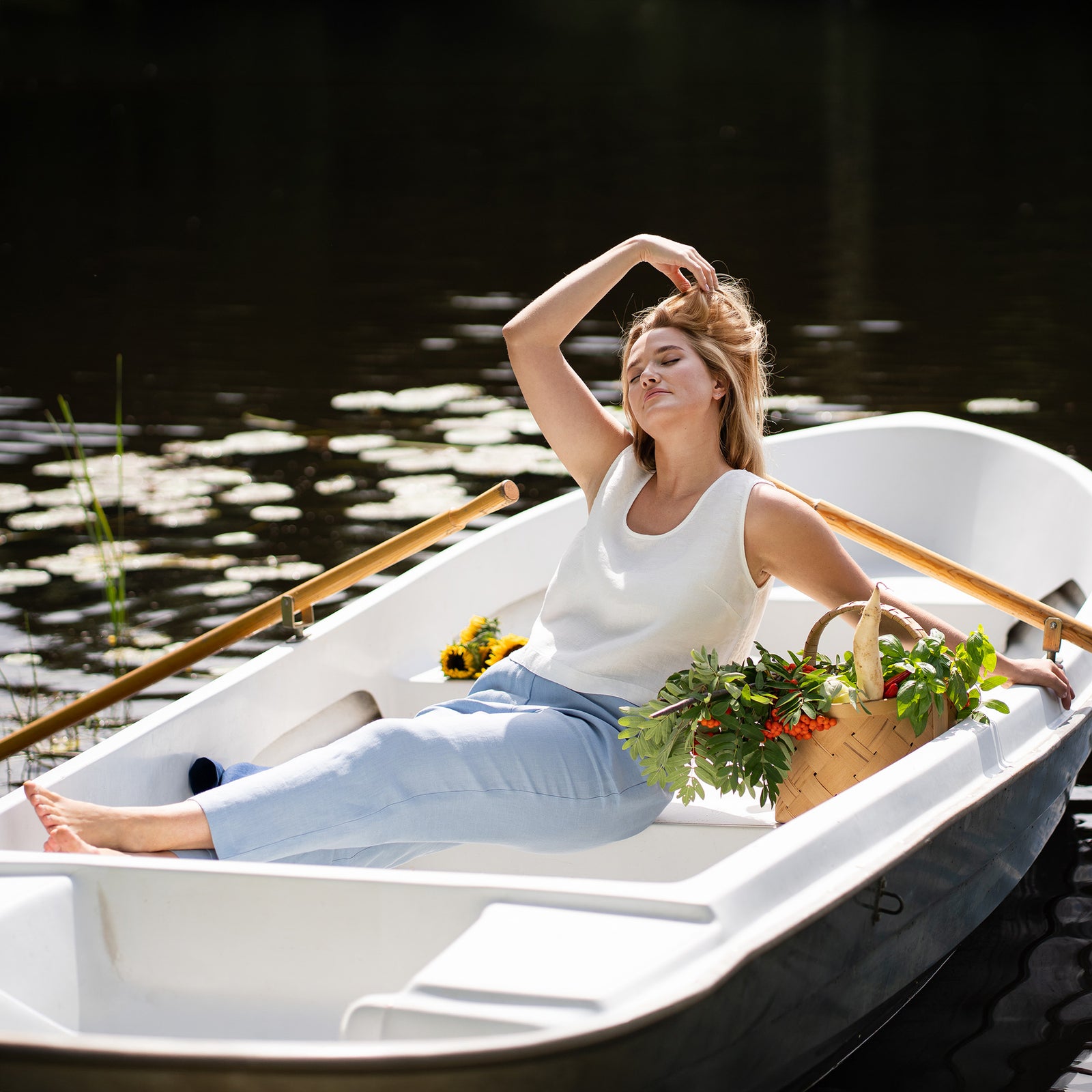 A woman is shown from the chest down wearing a white linen top and light blue linen trousers. She poses barefoot with her legs slightly crossed on a wooden dock beside a calm lake.