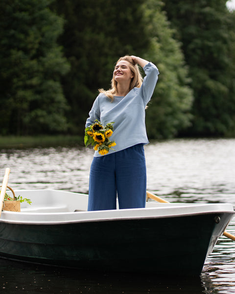 A woman stands in a small boat holding a bunch of sunflowers while running a hand through her hair and smiling warmly toward the sky. She is wearing a light blue linen shirt and dark blue pants.
