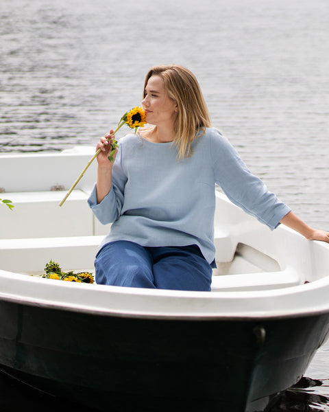 A woman sits inside a white rowboat on a calm lake, gently holding a bright yellow sunflower near her face. She wears a light blue linen top and dark blue pants, looking out over the water.