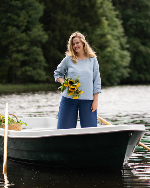 Standing up in a small white boat, a smiling woman holds a vibrant bouquet of sunflowers. She is dressed in a light blue, long-sleeved top and dark blue, wide-leg trousers, with a green shoreline behind her.