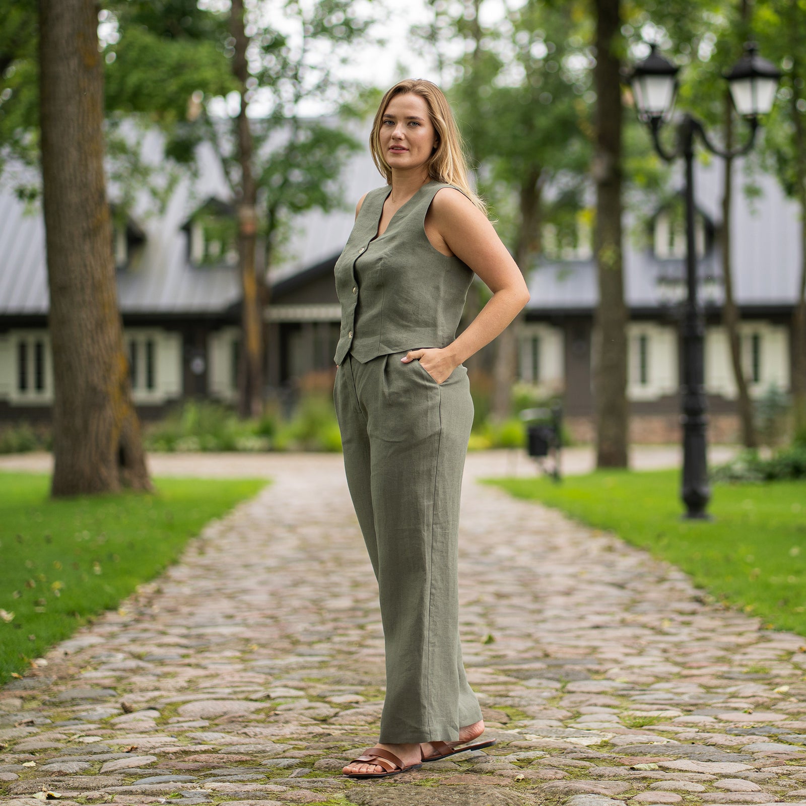 Een persoon wordt van de taille tot beneden getoond, die stenen groene, hooggetailleerde, linnen wijde broekspijpen draagt. Ze dragen zwarte sandalen met open tenen en dunne bandjes en hebben hun handen in de zij. De omgeving lijkt buiten te zijn op een asfaltpad, met een zwart metalen hek en groenblijvende planten op de achtergrond zichtbaar.