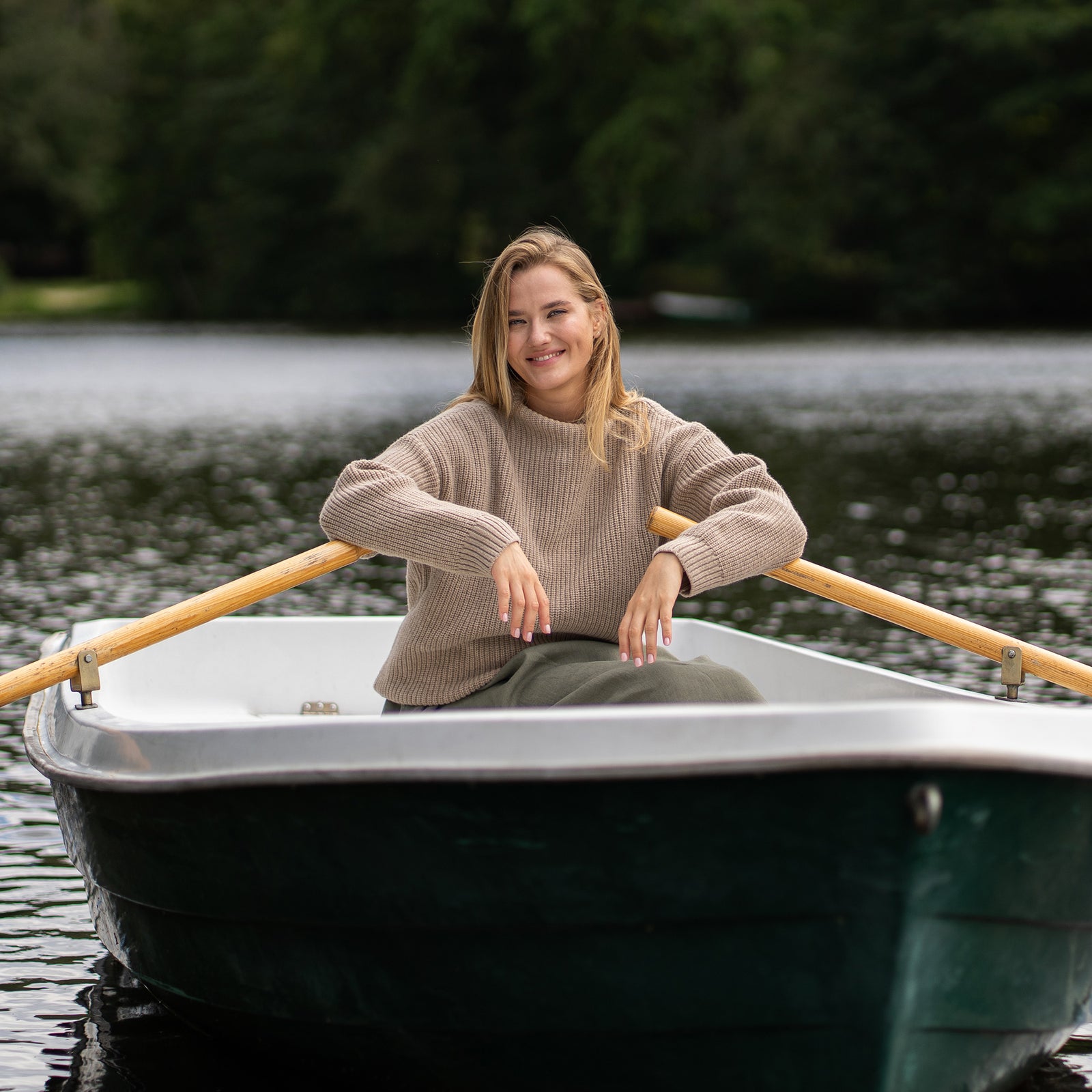 A woman relaxes in a small rowboat, wearing a beige mock-neck cashmere sweater over a stone green skirt. She is reaching into a woven basket full of fresh greens, with one hand casually resting on the wooden oar as she gazes thoughtfully over the water.