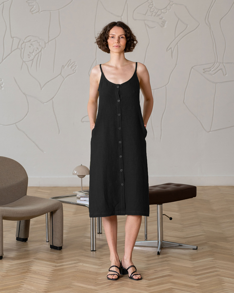 A woman with short, curly dark hair, wearing a sleeveless, button-front, black midi-dress with pockets and black open-toed sandals, stands with her hands in her pockets, looking directly at the viewer. Behind her is a light-colored wall with white raised line art depicting abstract figures. 