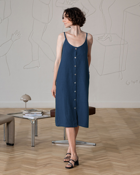 A woman with short, curly dark hair, wearing a sleeveless, button-front, storm blue midi-dress and black block-heeled sandals, stands facing slightly to her right, looking off into the distance. Her hands are relaxed at her sides.