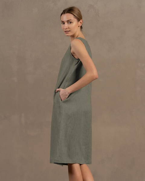 A woman with light skin and her hair styled back is shown in profile, facing slightly away from the viewer, wearing a sleeveless, stone green linen dress and resting one hand in the dress's side pocket.