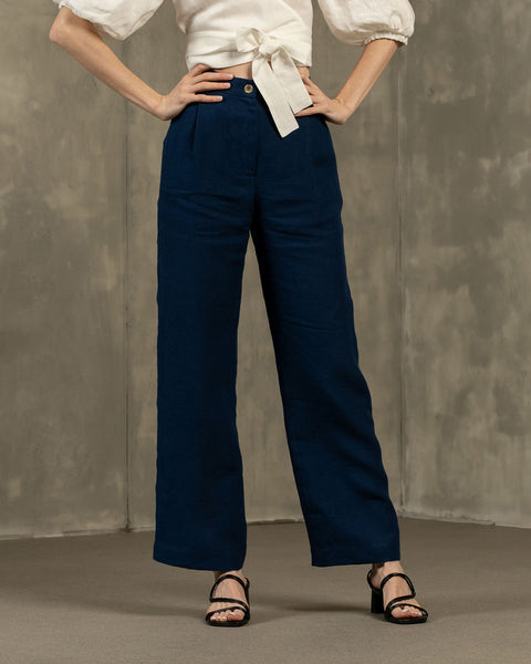A person is shown from the waist down, modeling menique, high-waisted, wide-leg linen pants. They have their hands on their hips and are wearing black open-toe heeled sandals. The background is a textured gray wall and floor.