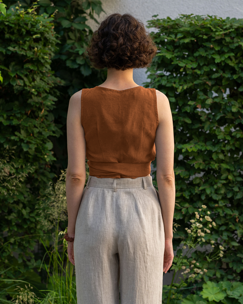 From the back, a light-skinned person with short, curly brown hair is standing outdoors. They are wearing an almond brown sleeveless wrap top and menique  high-waisted pants. Lush green foliage is visible in the background.