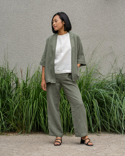 A woman with short dark hair smiles and looks to the right, wearing linen jacket, stone green wide-leg menique pants and black sandals. She is standing on a paved surface in front of a textured gray wall with tall green plants growing along the base.