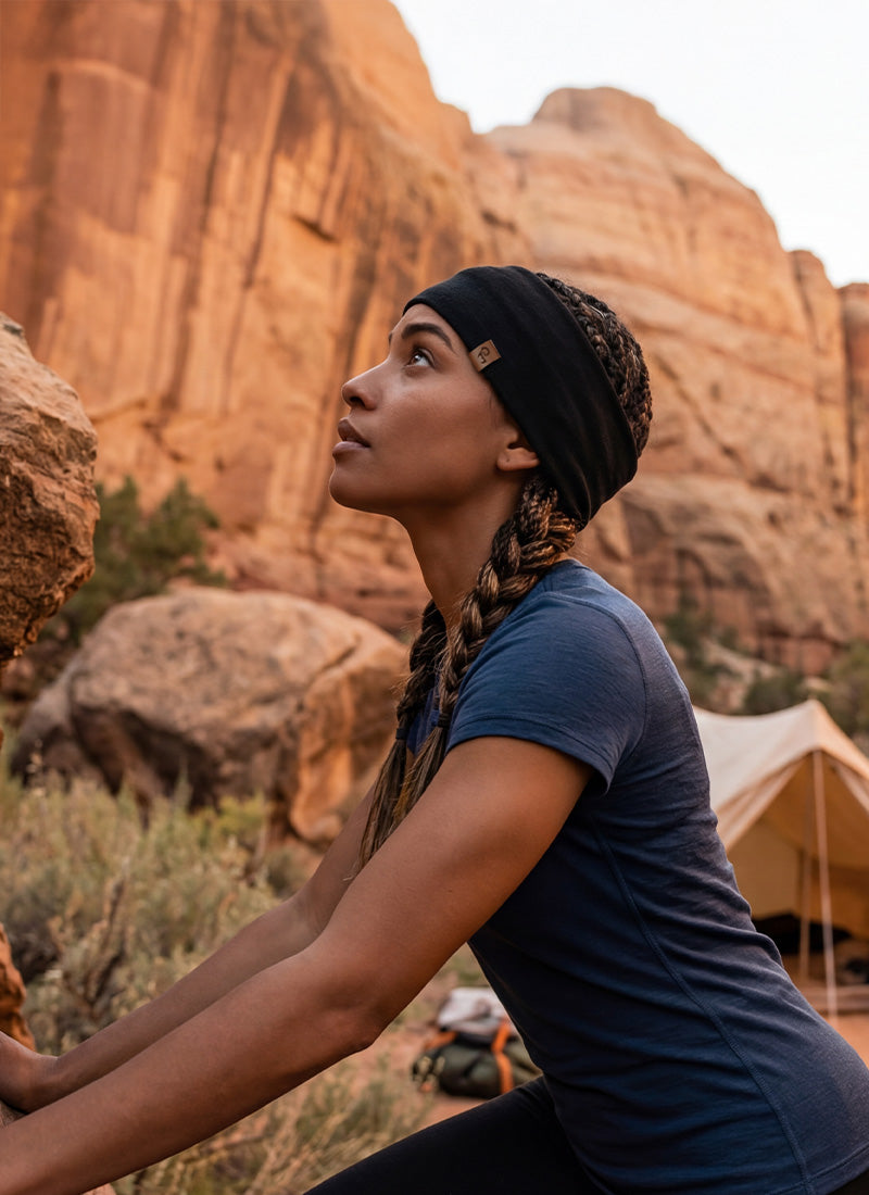 A side profile of a woman looking upwards as she leans against a large, textured red rock face. She has her hair in braids and is wearing a dark blue short-sleeved t-shirt and a wide black headband. A canvas tent is partially visible in the blurred background against the canyon walls.