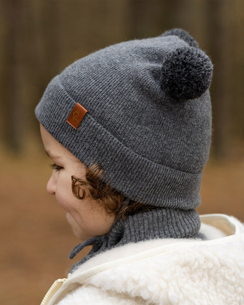 A young child with light brown curly hair is shown in profile, looking to the left. They are wearing a dark grey knit beanie with two dark grey pom-poms on top and a brown tag on the side, a grey turtleneck, and a cream-colored sherpa jacket. The background is a blurred forest.
