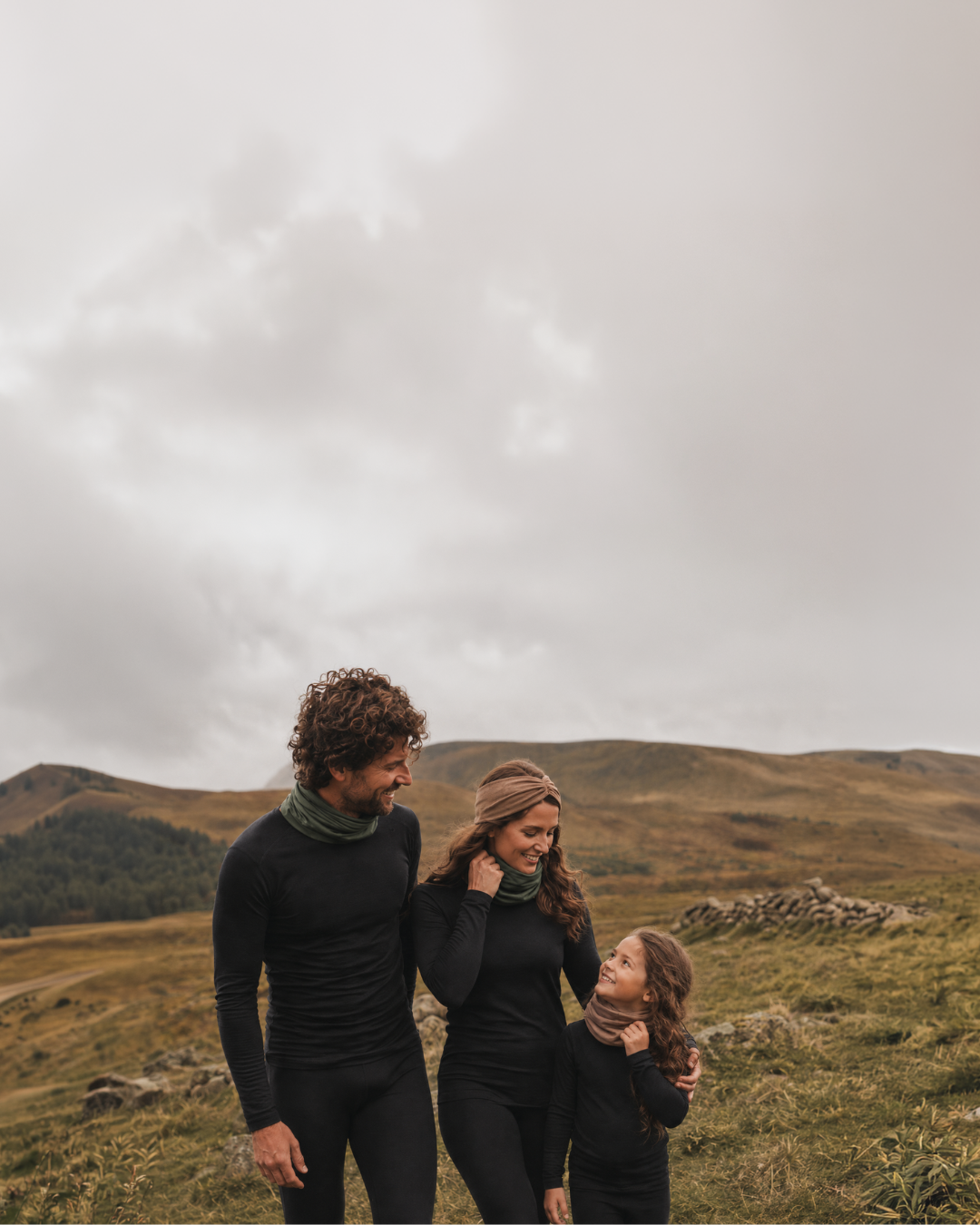 Three people wearing matching black activewear sets and scarves stand together in an open grassy landscape under a cloudy sky.