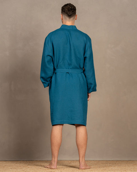 A man standing barefoot on a rug, wearing the menique cobalt blue men's linen bathrobe. There is a visible tie string at the back. The robe is knee-length.