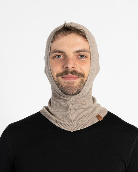 A man with a beard and mustache wears a creamy beige, fitted menique balaclava that covers his head and neck, with an opening for his face. He is also wearing a black long-sleeved shirt and is looking directly at the viewer with a slight smile.