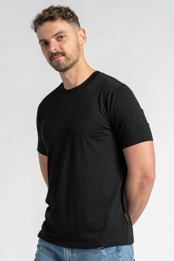 Een man die een zwart t-shirt van merinowol en een blauwe spijkerbroek draagt, is van borsthoogte te zien tegen een lichte achtergrond. Hij heeft kort bruin haar en een lichte baard en kijkt iets opzij. Het zwarte t-shirt van menique staat centraal en toont de pasvorm en het uiterlijk.