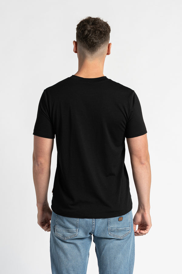 Achteraanzicht van een man die een zwart T-shirt en lichtblauwe spijkerbroek draagt, staand tegen een witte achtergrond. Het zwarte T-shirt staat centraal en toont de normale pasvorm en het ontwerp van achteren. De man staat in het midden, waardoor de achterkant van het kledingstuk duidelijk zichtbaar is.
