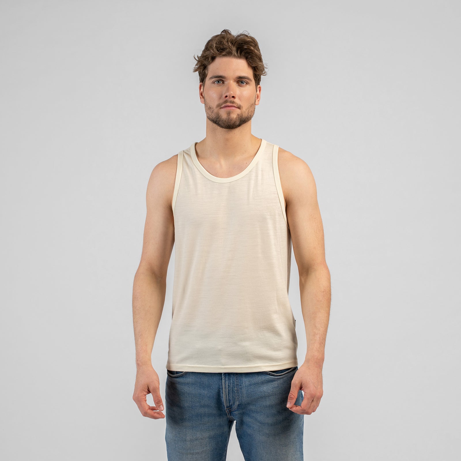 A studio portrait of a man standing and looking back over his right shoulder. He is wearing a light beige tank top and medium-wash blue jeans, with his left hand resting in his front pocket. The background is a solid light grey.