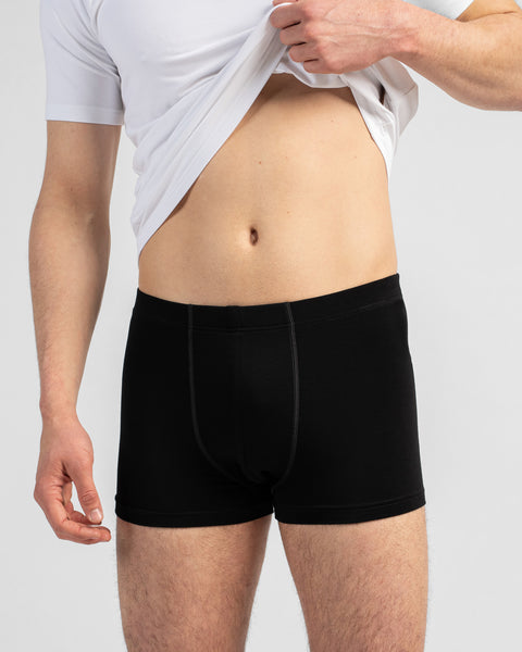 A close-up shot of a man's torso as he pulls up a white t-shirt to reveal his abdomen and black boxer briefs. The background is a plain light grey.