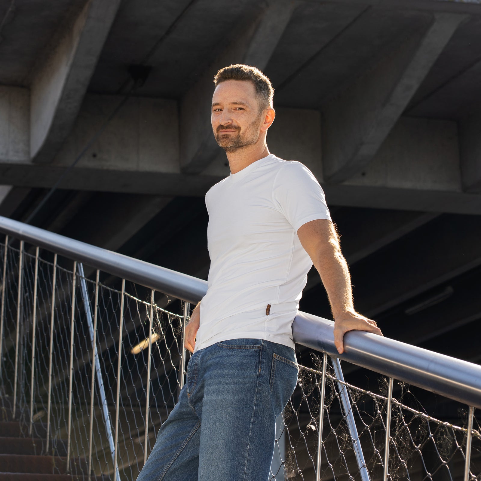 A man stands outdoors against a backdrop of rusted steel beams and a modern concrete overpass. He is wearing a plain white short-sleeved t-shirt and blue jeans. His right hand rests on his hip, and his left hand is tucked into his front pocket as he looks off into the distance.