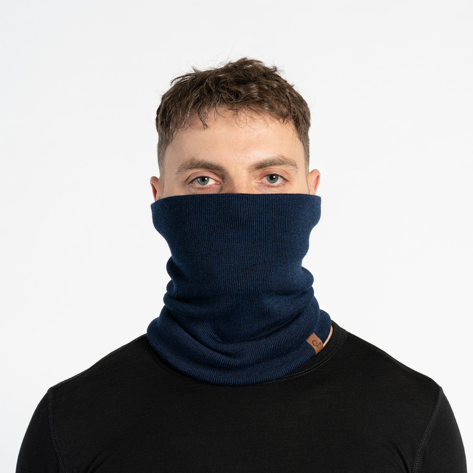 A person with short brown hair is shown in profile, wearing a black knit neck gaiter pulled up to cover their nose and mouth. They are wearing a blue long-sleeved shirt. The background is a plain light gray. A small brown tag is visible on the neck gaiter.