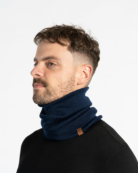 A person with short brown hair and a mustache is shown in a studio setting against a white background, wearing blue knit neck gaiter. They are also wearing a black long-sleeved shirt. The neck gaiter has a small brown tag. The person is looking slightly to their left.