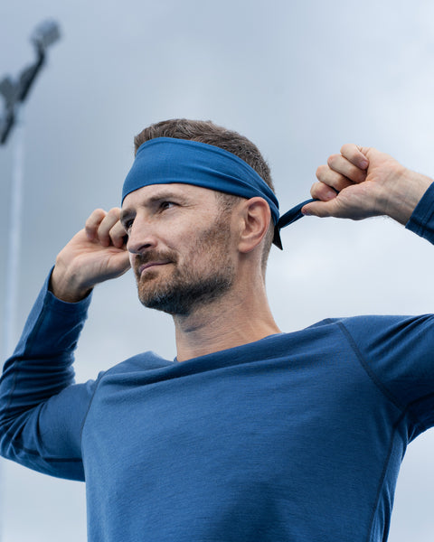 A low-angle shot of a man outdoors tying a blue athletic headband behind his head. He is looking off to the side with a focused expression, wearing a matching blue long-sleeve shirt. The background features a cloudy sky and a blurred, tall stadium light pole.
