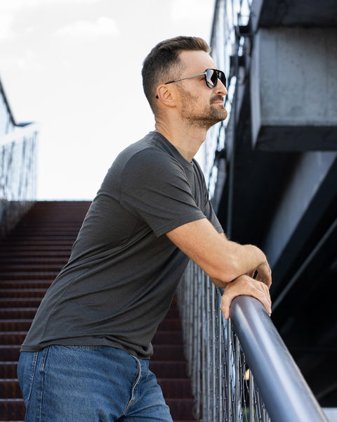 A man wearing sunglasses stands on outdoor metal stairs, leaning forward with both hands resting on the handrail and looking off to the right. He is wearing a charcoal grey short-sleeve crew neck t-shirt and blue jeans. The background features rusted metal steps and a dark industrial concrete and steel structure against a bright sky.