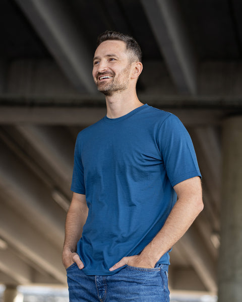 A smiling man stands outdoors with his hands in his jeans pockets, looking happily upward and to the left. He is wearing a short-sleeve, denim-blue crew neck t-shirt. The background features the underside of a concrete overpass with angled beams.