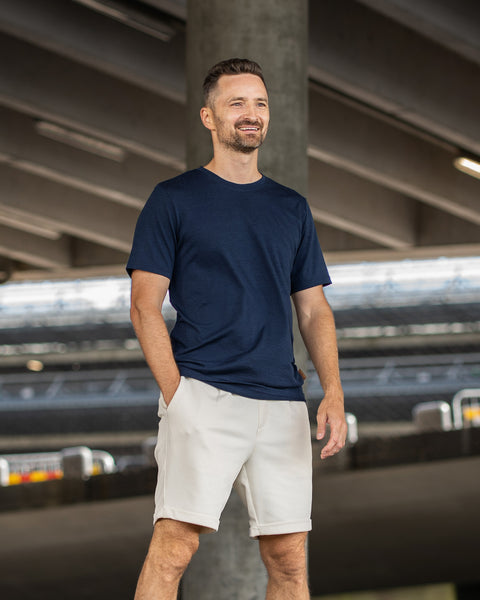 A smiling man with a short beard stands looking off to the right, with one hand resting in his pocket. He is wearing a navy blue short-sleeve crew neck t-shirt and light beige shorts. The background features the shaded underside of a large concrete structure or overpass.