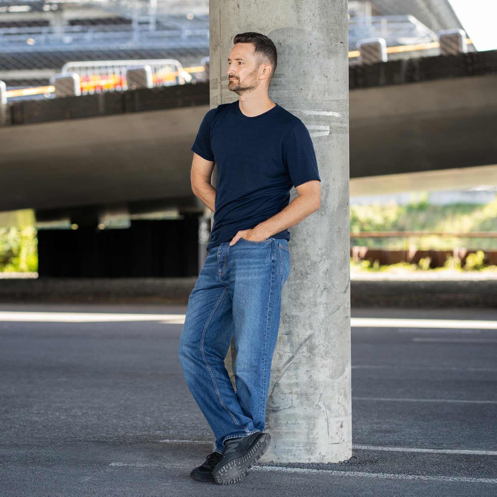 A man with a short beard smiles and looks to the right while standing with both hands tucked into the front pockets of his blue jeans. He is wearing a navy blue short-sleeve crew neck t-shirt. The background is a shaded outdoor area beneath a concrete overpass.