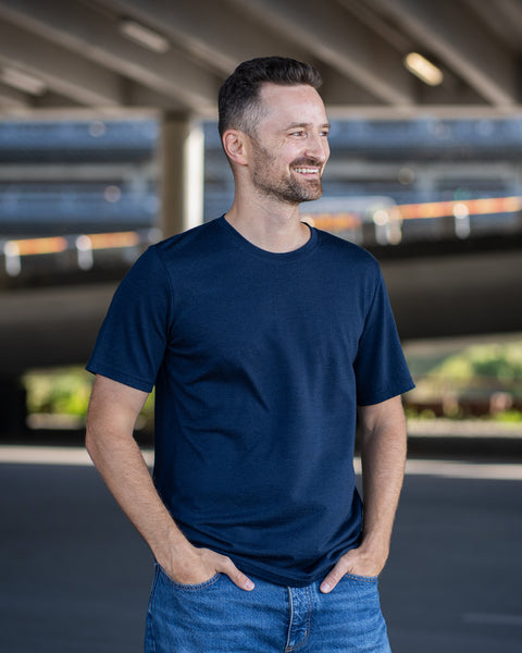 A man with a short beard smiles and looks to the right while standing with both hands tucked into the front pockets of his blue jeans. He is wearing a navy blue short-sleeve crew neck t-shirt. The background is a shaded outdoor area beneath a concrete overpass.