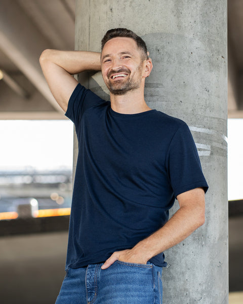 A laughing man leans casually against a large, round concrete pillar. He has one hand tucked into his blue jeans pocket and the other arm raised, resting comfortably behind his head. He is wearing a navy blue short-sleeve crew neck t-shirt, set against the blurred backdrop of a concrete underpass.