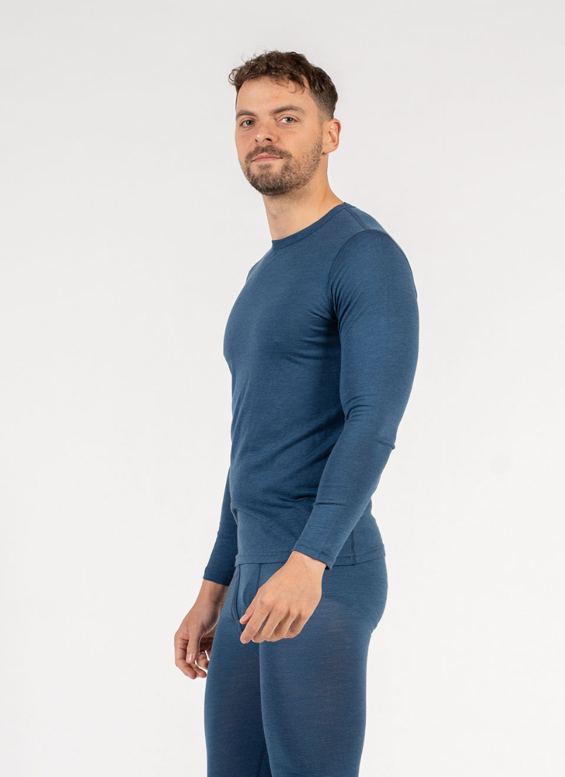 A man is shown from a slight side angle wearing a form-fitting blue long-sleeve base layer top and matching blue leggings. He has a short beard and is looking directly at the camera against a plain white backdrop.