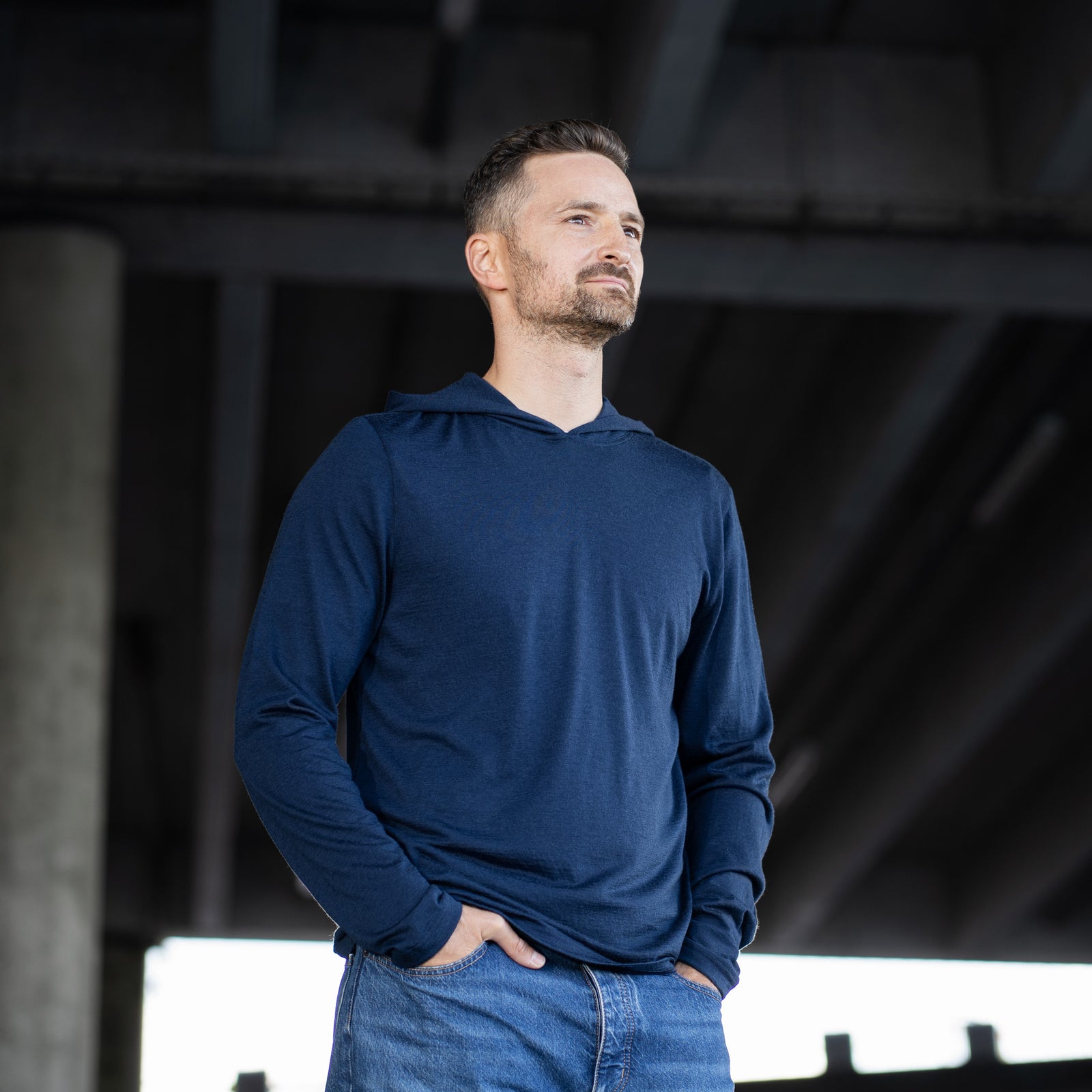 A smiling man stands outdoors with both hands in the front pockets of his blue jeans. He is wearing a long-sleeve, navy blue hoodie. The background shows the underside of a large concrete overpass structure.