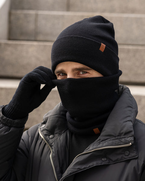 Man wearing a black Merino wool beanie, gloves, and neck gaiter, partially covering his face, styled with a black puffer jacket on outdoor steps in winter.