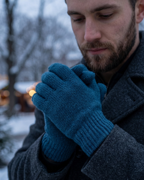 A man standing outdoors in a snowy setting, looking down gently at his clasped hands. He is wearing a dark gray coat and thick, dark blue knitted wool gloves. The background shows blurred winter trees and warm, glowing string lights.
