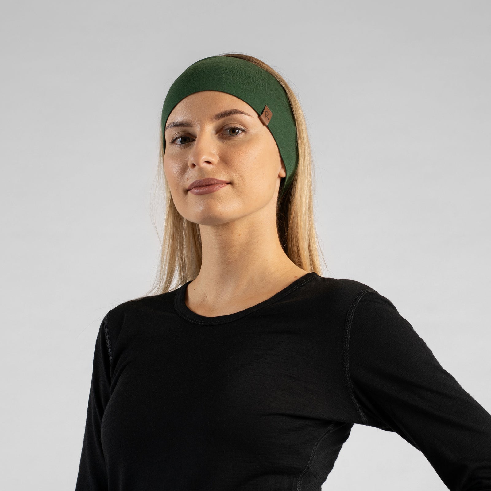 De foto toont een vrouw die in een bos zit, gekleed in de uniseks donkergroene menique merino HOEDEBAND. Ze draagt ook een mouwloos zwart merino hemd. Ze kijkt naar de zijkant.