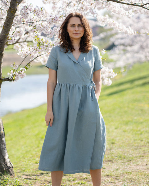A woman with brown hair, wearing a light blue, short-sleeved linen wrap dress, stands outdoors next to a tree with white blossoms.
