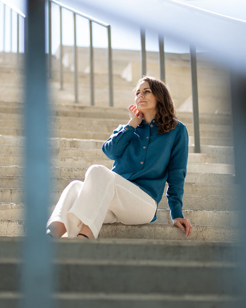 A person with long, dark, curly hair sits casually on light concrete steps. They are wearing a teal long-sleeved collared shirt, white wide-leg pants, and dark sandals. A metal railing is visible on the right side of the frame.